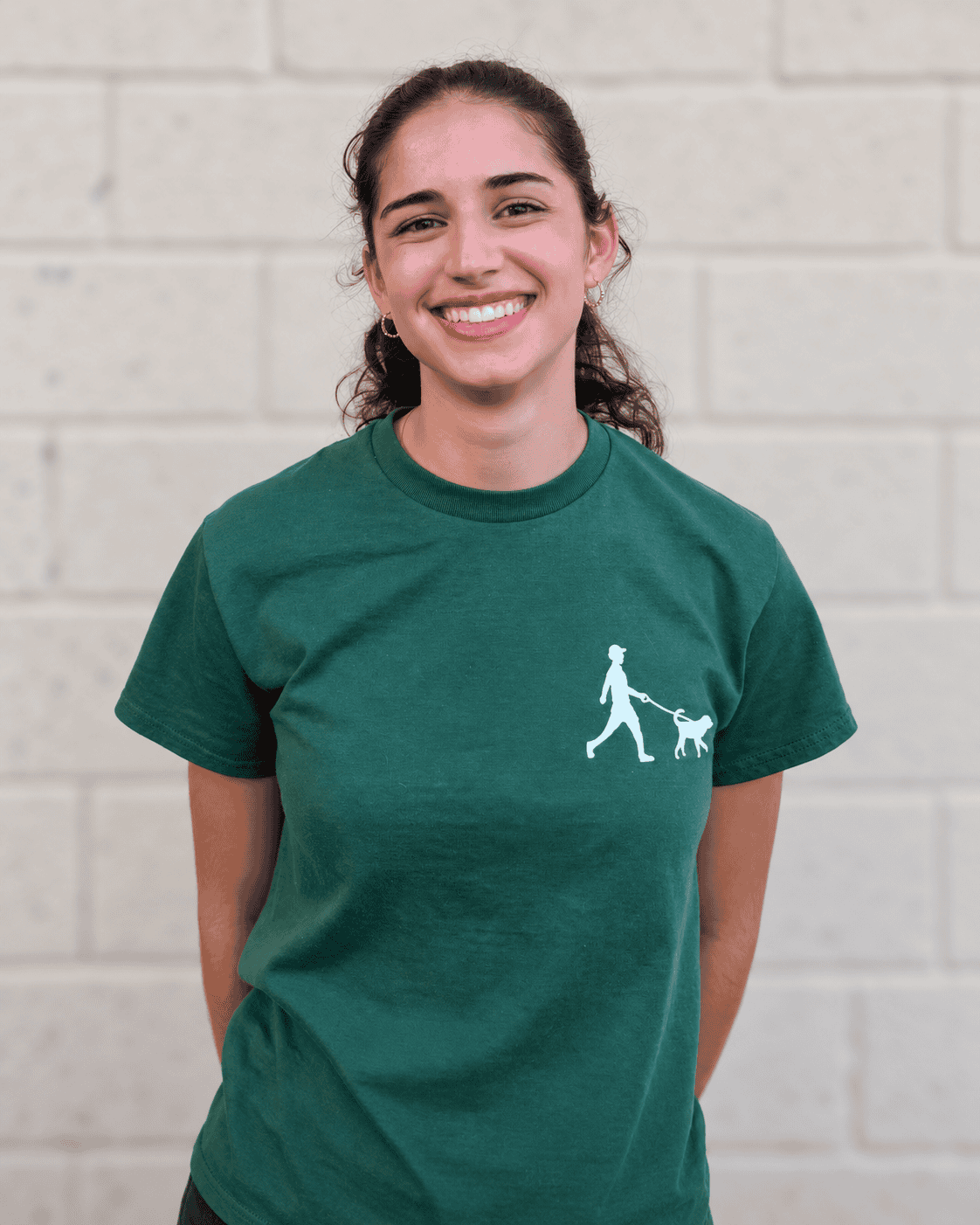 Smiling Bethesda Dog Walkers team member wearing a green company T-shirt in front of a light-colored block wall.