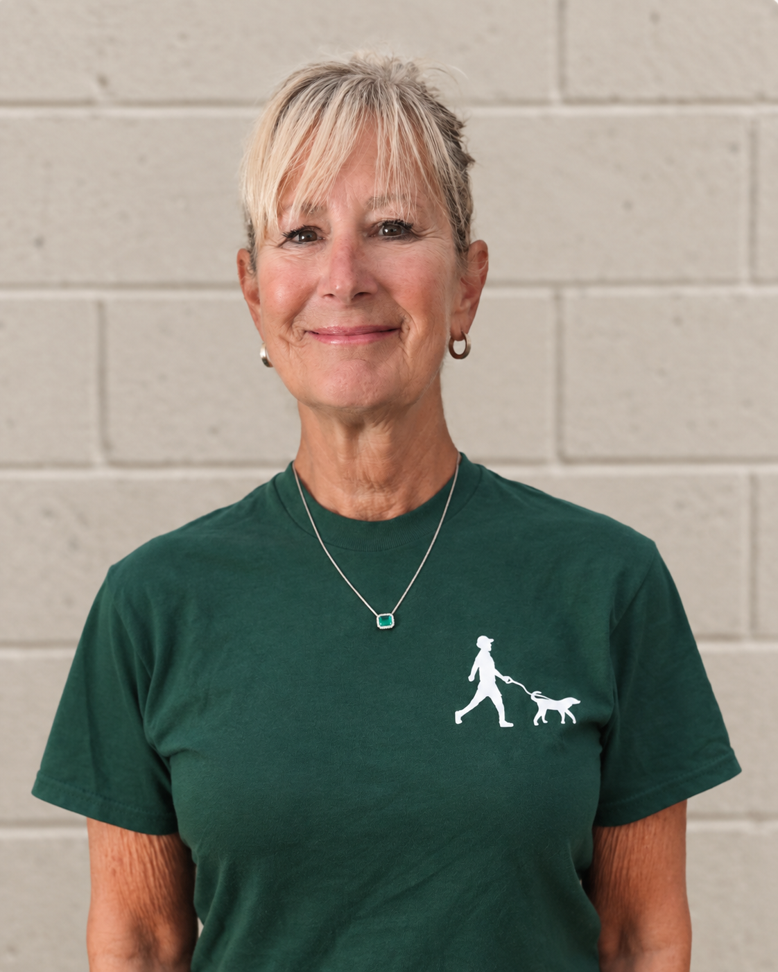 Emily Shaw, Bethesda Dog Walkers team member, smiling in a green company shirt against a beige brick wall.